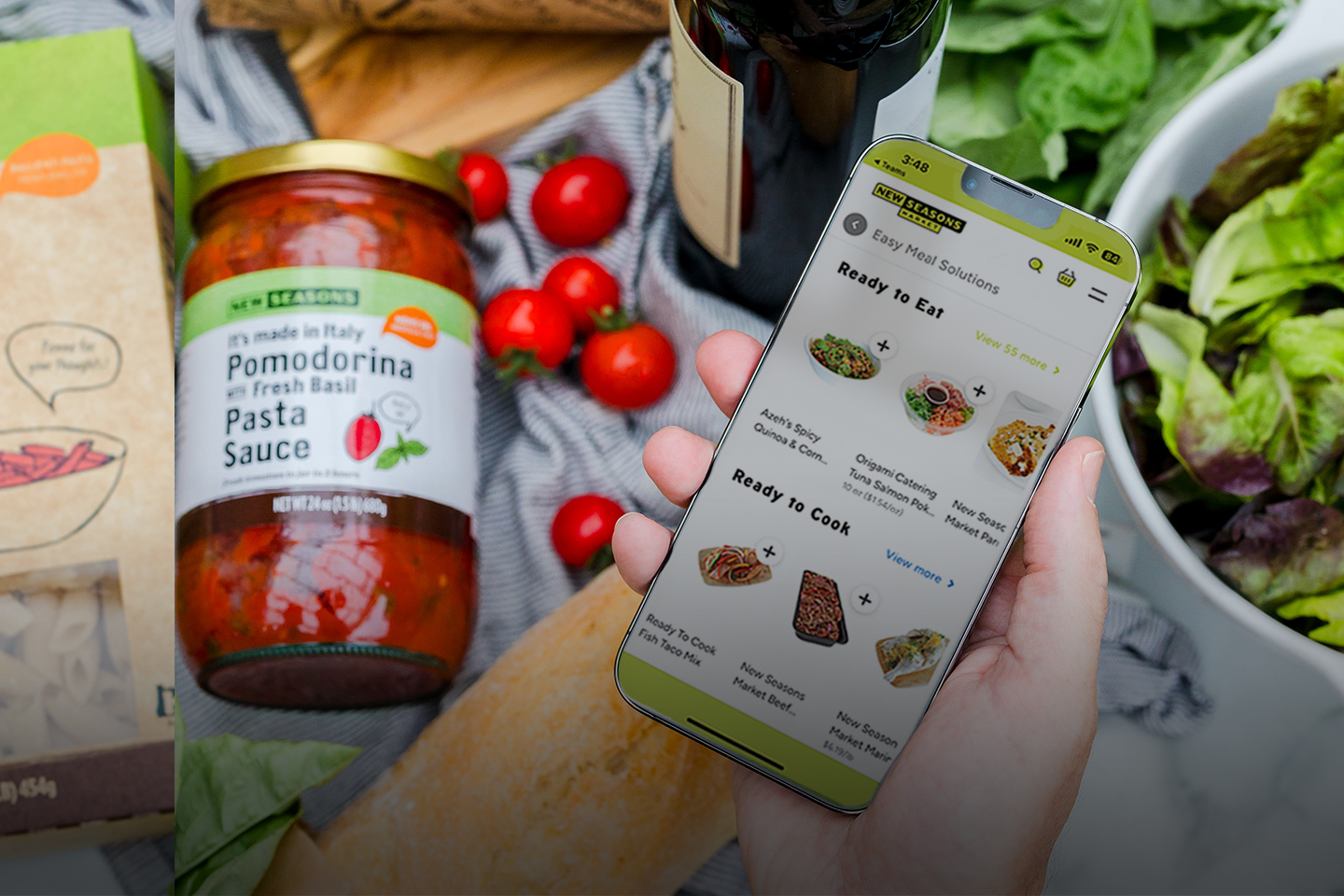 Hand holding a phone shopping online with New Seasons above an array of grocery items like pasta and a jar of sauce.