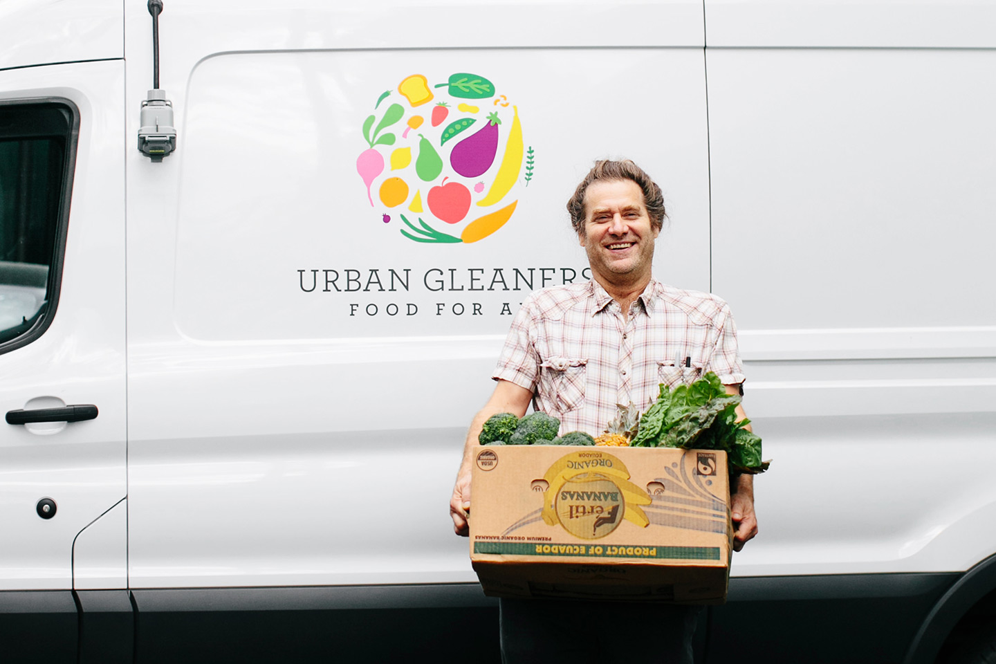 Person holding box of fresh vegetables in front of an Urban Gleaner white van.