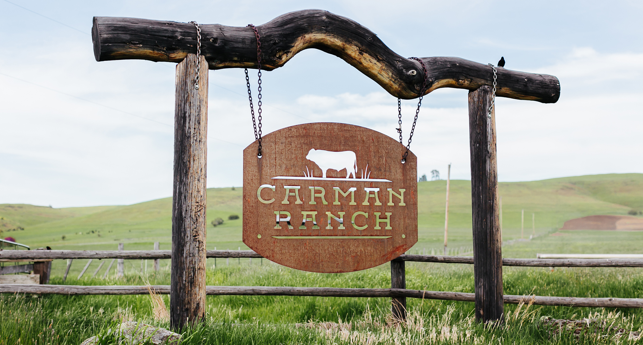Metal Carman Ranch sign hangs on a rustic wooden frame at farm entrance.