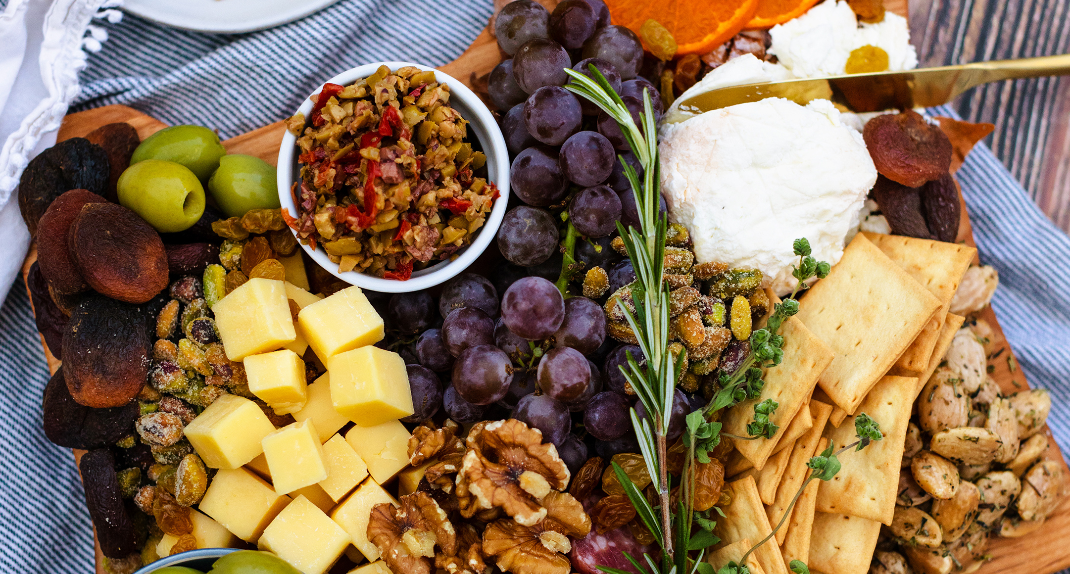 a bountiful cheese board with fruit, nuts, crackers and multiple cheeses on a wooden board set on a blue towel